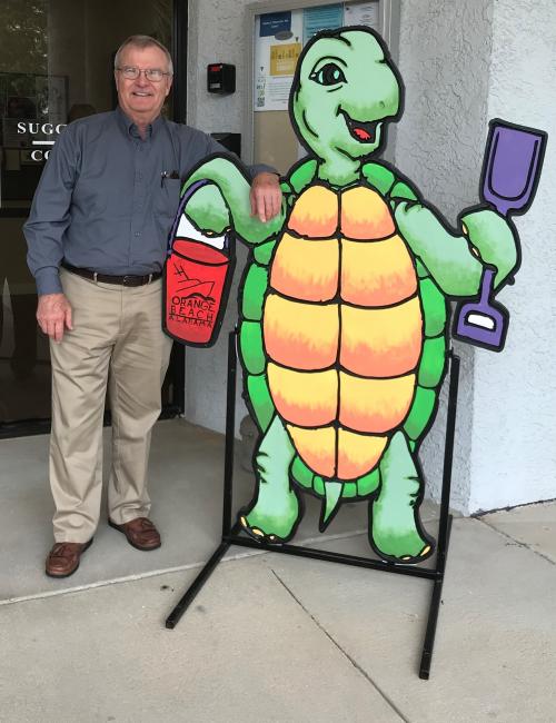 Photo of Dale Barstow next to a painted wooden cut out of a turtle holding shovel and sand bucket.