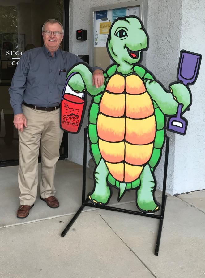 Photo of Dale Barstow next to a painted wooden cut out of a turtle holding shovel and sand bucket.